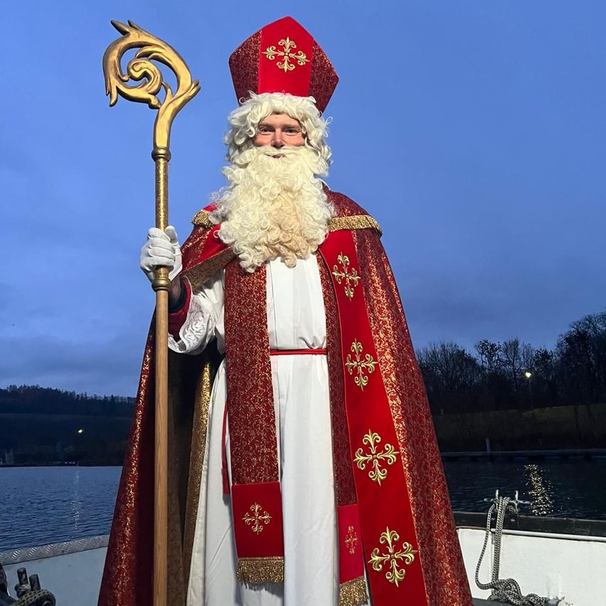 Eine Person in Bischofskleidung mit rotem Hut und Robe, die einen goldenen Stab hält, steht auf einem Boot in der Nähe eines Gewässers.