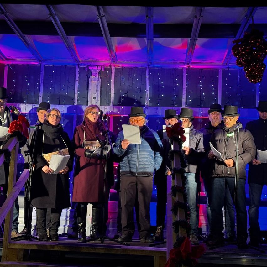 Eine Gruppe von Menschen steht auf der Bühne und singt mit Mikrofonen. Sie sind in Winterkleidung gekleidet und einige tragen Hüte. Hinter ihnen befinden sich rote Blumen und Dekorationen.