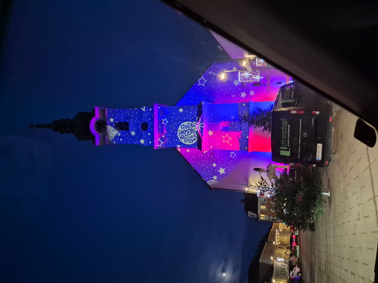 Ein Gebäude mit Sternen- und Kreismustern ist nachts mit blauen und lila Lichtern beleuchtet. Das Gebäude hat eine Uhr, und ein Van ist davor geparkt.