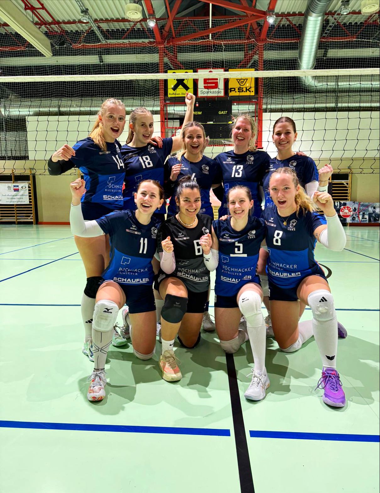 Eine Volleyballmannschaft posiert für ein Foto mit erhobenen Händen, trägt blaue Trikots mit Nummern und weißen Socken, mit einem Volleyballnetz im Hintergrund.