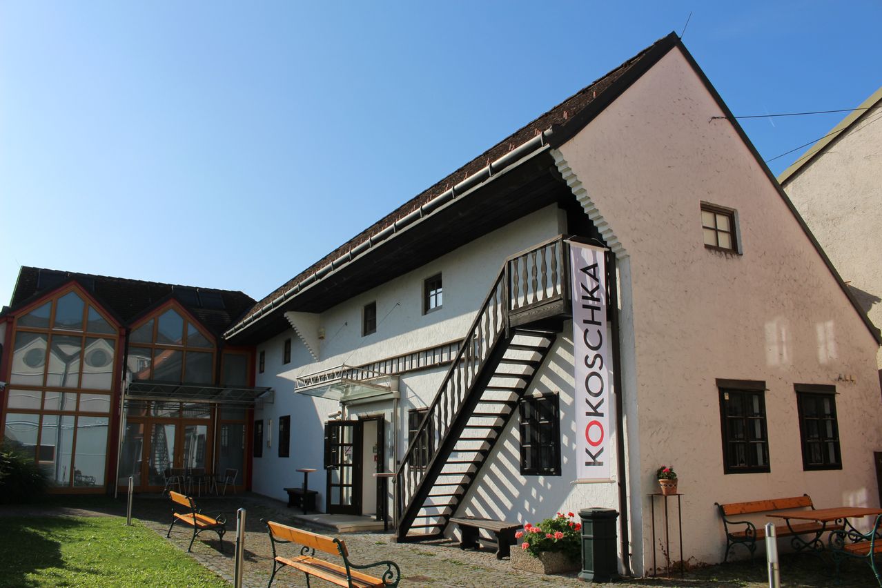 Ein weißes Gebäude mit einem braunen Dach, das eine Treppe mit einer Metallbrüstung aufweist. An der Wand hängt ein Banner mit dem Wort 'KOKOSCHKA'. Vor dem Gebäude befinden sich Bänke und ein Mülleimer.