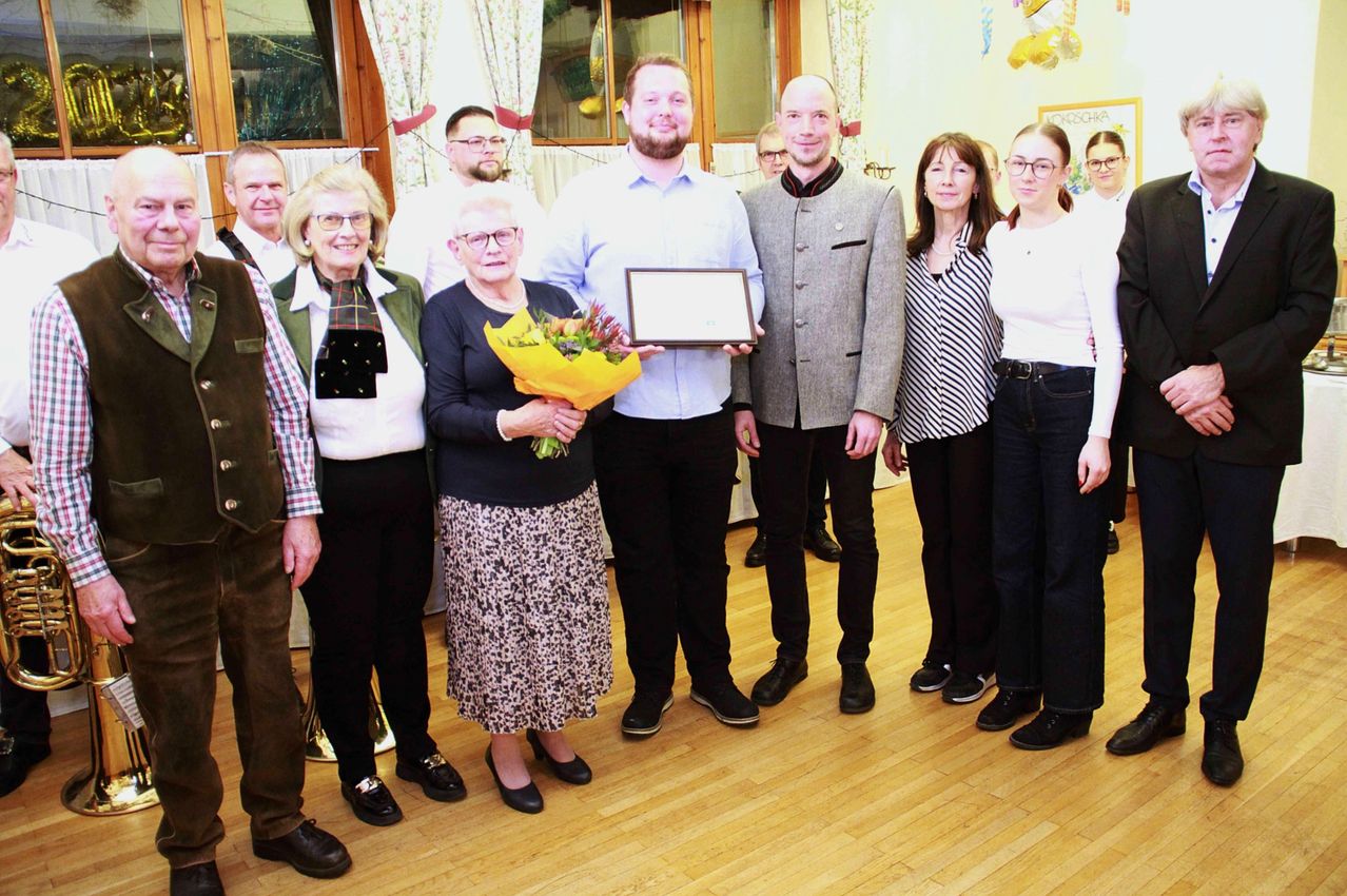 Mehrere Menschen stehen auf einem Holzboden, wahrscheinlich in einem Sitzungssaal. Zwei Männer halten ein Zertifikat, während eine Frau in der Mitte Blumen hält. Hinter ihnen lächeln zwei Frauen und ein Mann.