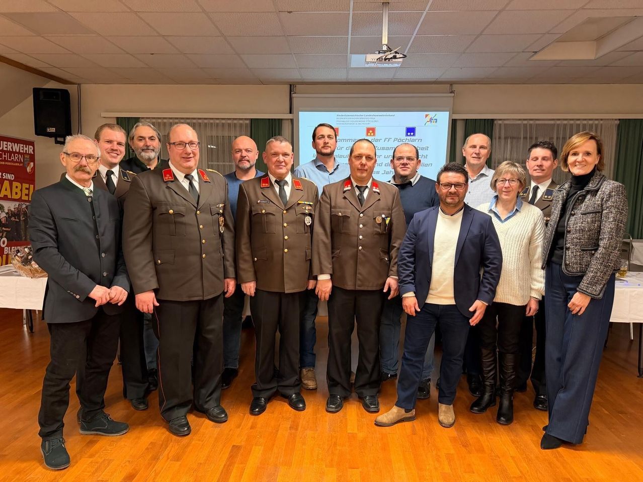 Eine Gruppe von Personen, einige in Militäruniformen, steht in einem Raum mit Holzböden. Vor ihnen befindet sich eine Projektionsleinwand mit angezeigtem Text.