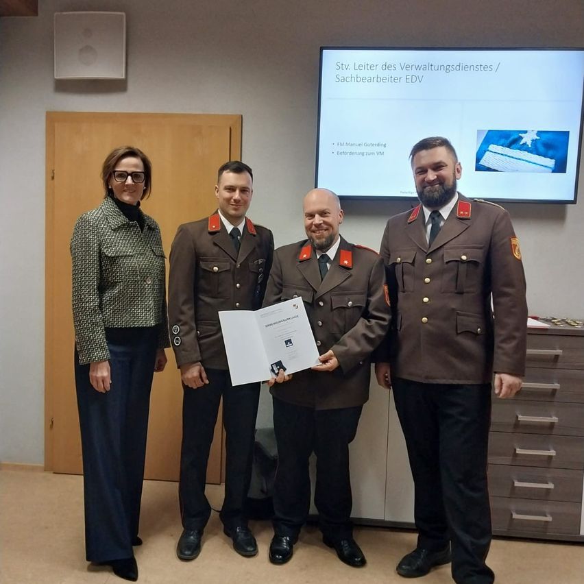 Vier Personen stehen in einem Büro, mit einer Frau auf der linken Seite und drei Männern in Militäruniformen auf der rechten Seite. Sie posieren für ein Foto mit einem Zertifikat. Ein Monitor ist an der Wand hinter ihnen montiert.