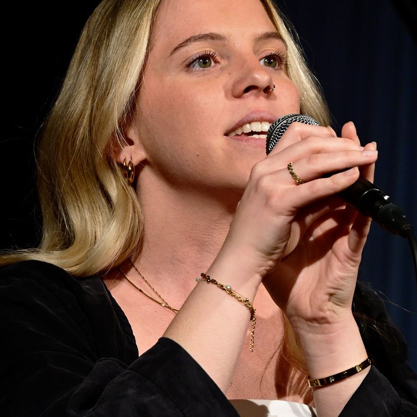 Eine Frau mit blonden Haaren, die ein schwarzes Oberteil trägt, singt in ein Mikrofon. Sie hat einen goldenen Ring, Armband und eine Halskette an.