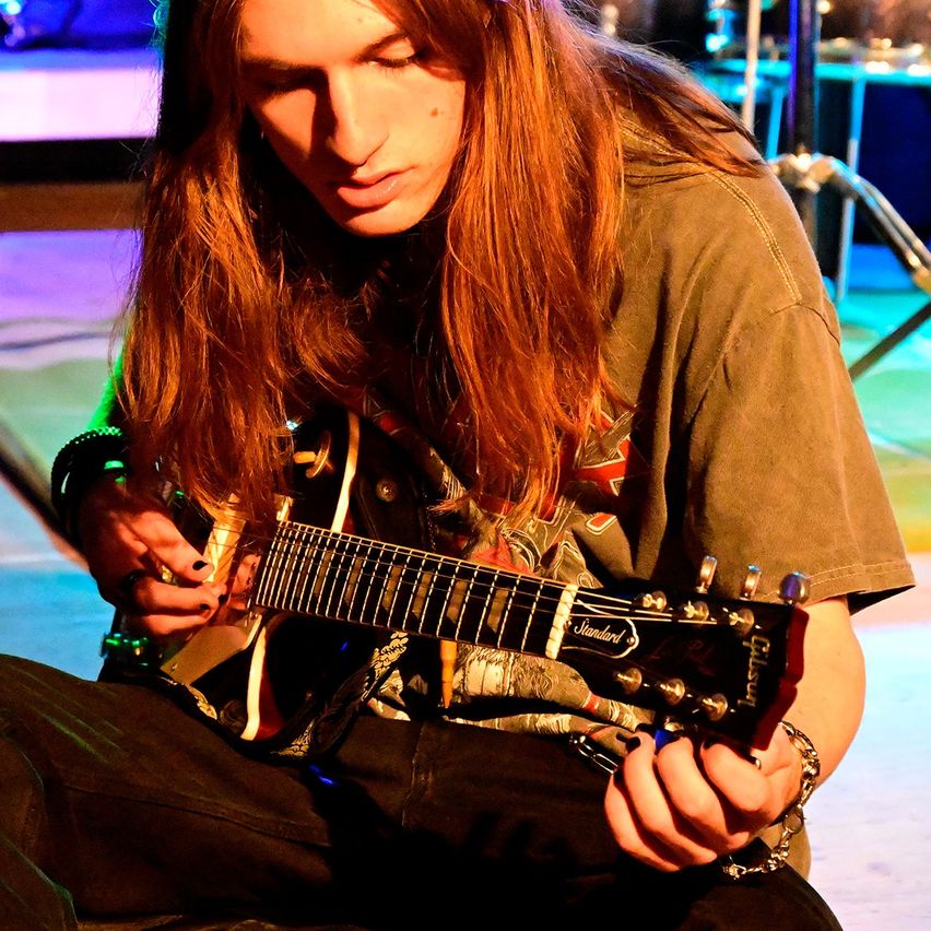Eine Person mit langen Haaren sitzt und spielt eine Gitarre. Die Gitarre hat das Wort 'Gibson' darauf. Die Person trägt ein braunes Hemd und Hose. Der Hintergrund ist unscharf.