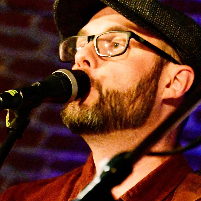 Ein bärtiger Mann mit Brille singt in ein Mikrofon auf der Bühne. Er trägt ein braunes Hemd und einen schwarzen Fedora.