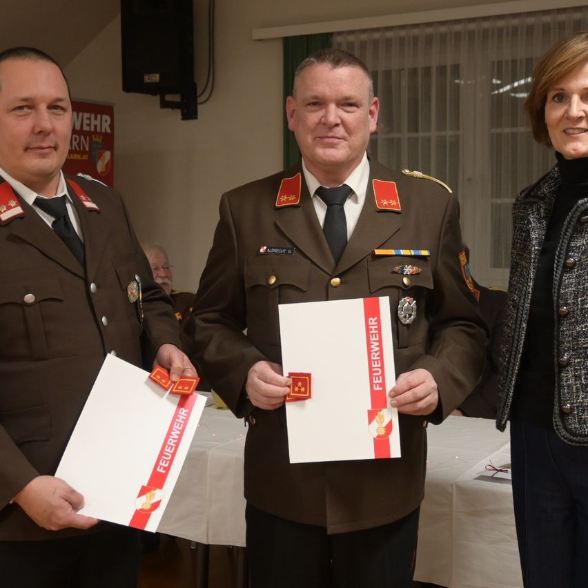 Drei Personen in Uniformen halten Zertifikate. Der Mann in der Mitte trägt eine rote Sternabzeichen. Hinter ihnen steht ein Tisch mit weißer Tischdecke.