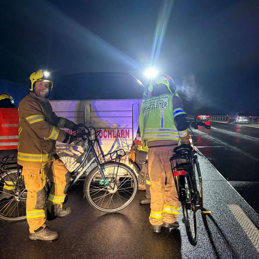 Zwei Feuerwehrleute stehen bei Nacht am Straßenrand, mit einem Feuerwehrwagen und Fahrrädern. Einer trägt eine reflektierende Weste und hält ein Fahrrad. Hinter ihnen fahren Autos auf der Straße.