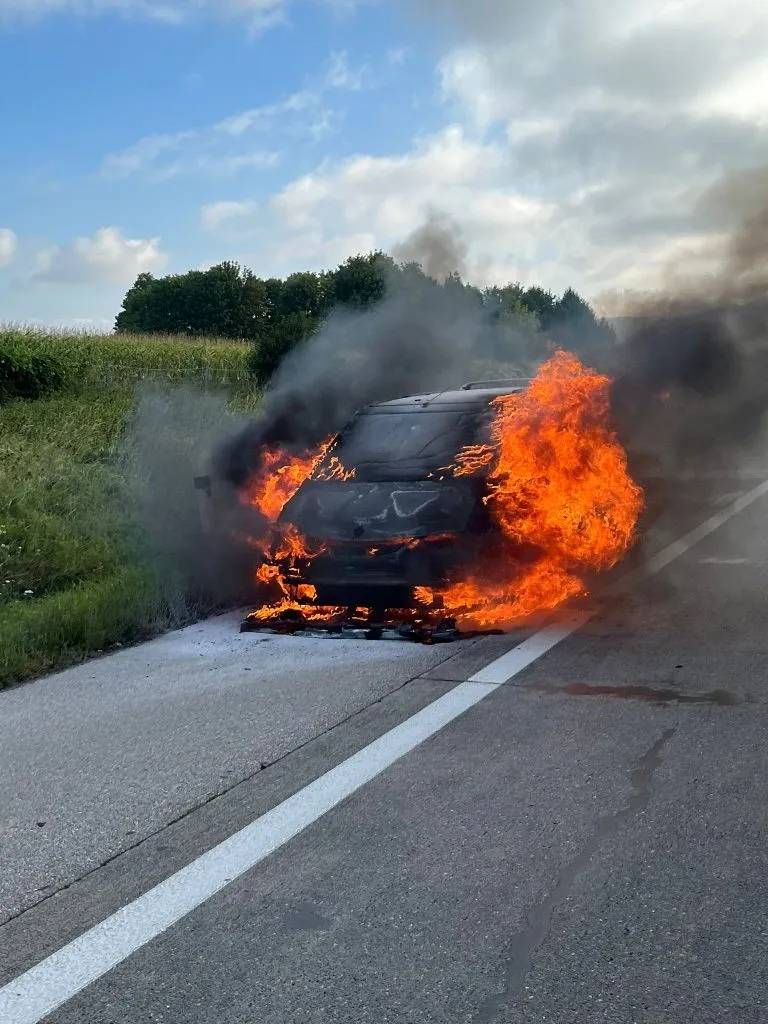 Ein brennendes Auto steht am Straßenrand und gibt dicken Rauch ab, wobei die Flammen bis zum Dach reichen.