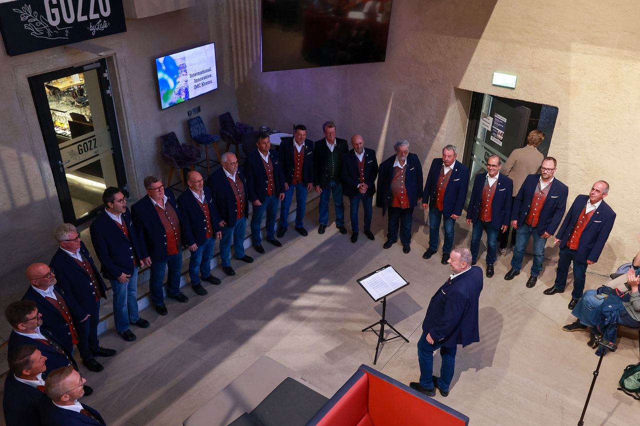 Ein Chor von Männern in formeller Kleidung, aufgestellt und singend in einem Raum mit einer Bühne. Ein Mann steht vor ihnen und leitet möglicherweise die Aufführung.