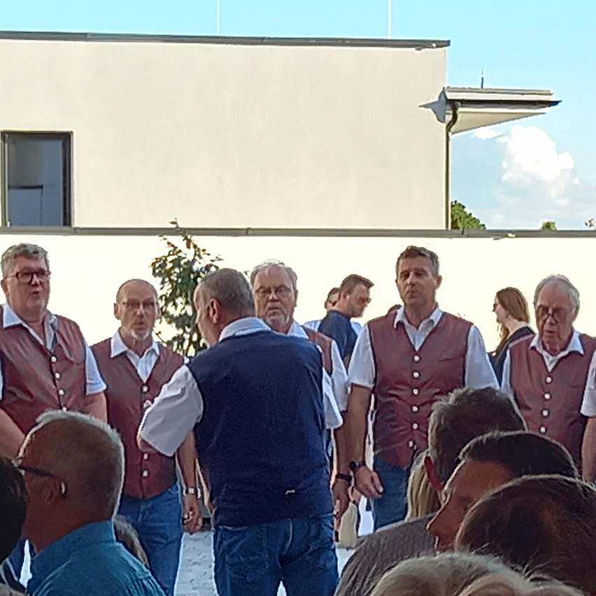 Eine Gruppe von Männern in Westen, einige in Weinrot und andere in Blau, stehen zusammen vor einem Gebäude. Sie scheinen in ein Gespräch vertieft zu sein.