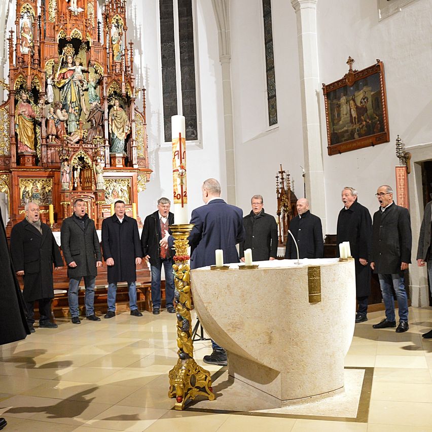 Ein Chor steht in einer Kirche vor einem Altar mit Kerzen. Sie tragen Mäntel und singen. Dahinter steht ein hölzerner Altar mit einer Statue der Jungfrau Maria.
