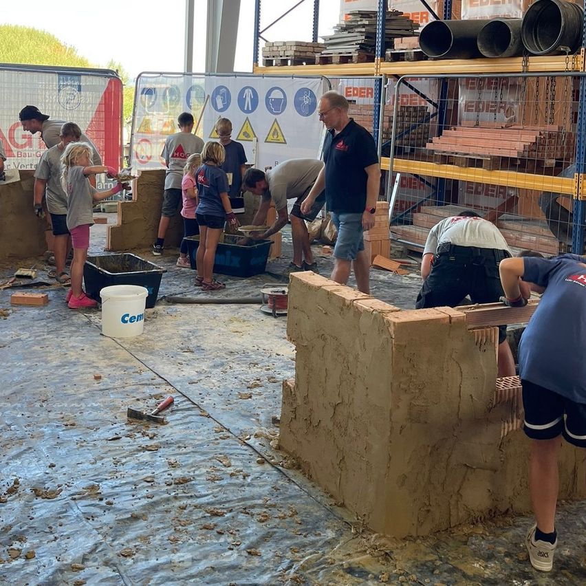 Mehrere Personen, einschließlich Kinder, arbeiten unter Aufsicht an einer Ziegelmauer. Sie verwenden Werkzeuge und Eimer mit Zement. Die Umgebung ist im Freien, mit Regalen und einem Zaun im Hintergrund.