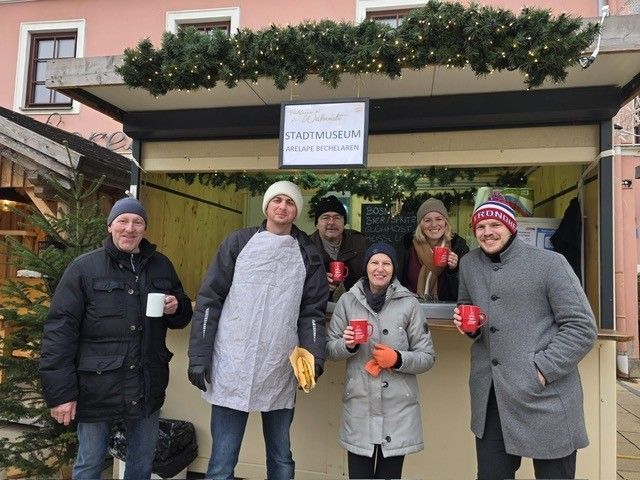 Eine Gruppe von Menschen in Winterkleidung posiert für ein Foto vor einem festlich geschmückten Stand. Sie halten rote Tassen und ein Schild darüber lautet 'Stadtmuseum Arelape Becheleraren.'