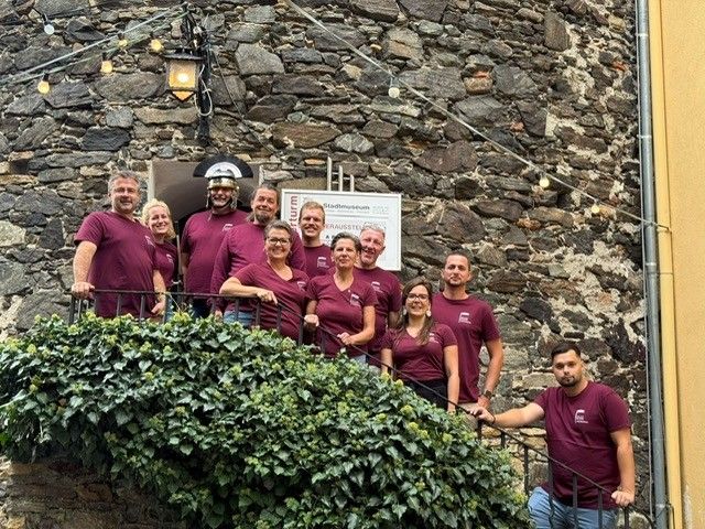 Ein Team von Personen in Weinrot-Shirts posiert für ein Foto vor einem Steingebäude mit einem Schild.