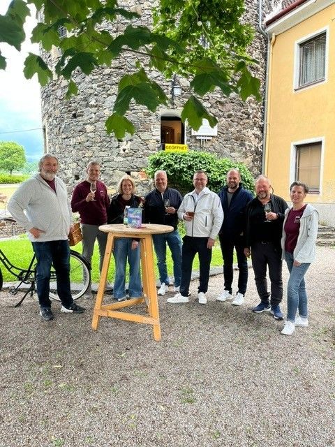 Eine Gruppe von Menschen posiert draußen in der Nähe eines kleinen Tisches, mit Weingläsern in der Hand, vor einem Gebäude mit einem Schild, das 'GEOFFNET' steht. Ein Fahrrad und einige Bäume sind in der Nähe.