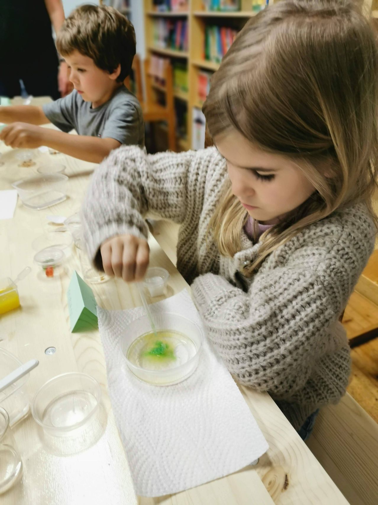 Ein kleines Mädchen führt ein Experiment an einem Tisch mit verschiedenen Labormaterialien durch. Sie hält eine Pipette und betrachtet eine Petrischale mit grüner Flüssigkeit.