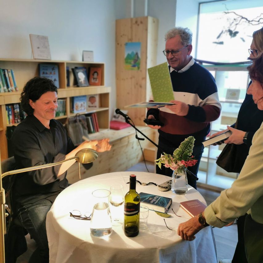 Eine Gruppe von Menschen ist in einem Raum mit einem Tisch. Ein Mann mit Brille hält ein Buch, und ein anderer Mann sitzt mit einem Buch. Eine Frau steht in der Nähe eines Tisches mit Büchern und Blumen.