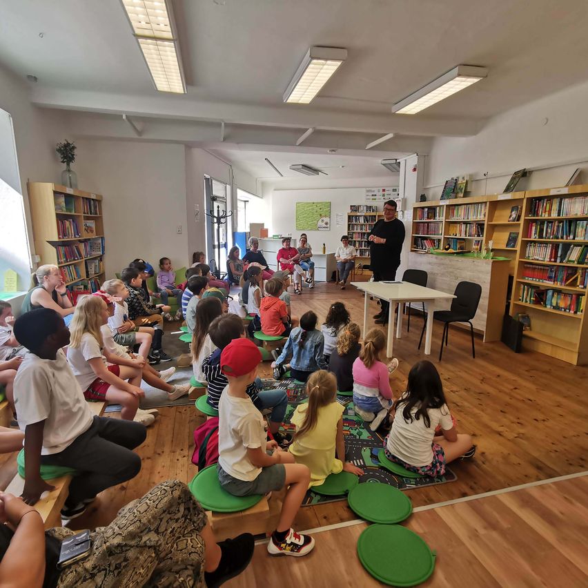 Eine Gruppe von Kindern und Erwachsenen sitzt in einer Bibliothek auf Stühlen und hört einem Mann zu, der vor einem Tisch steht.