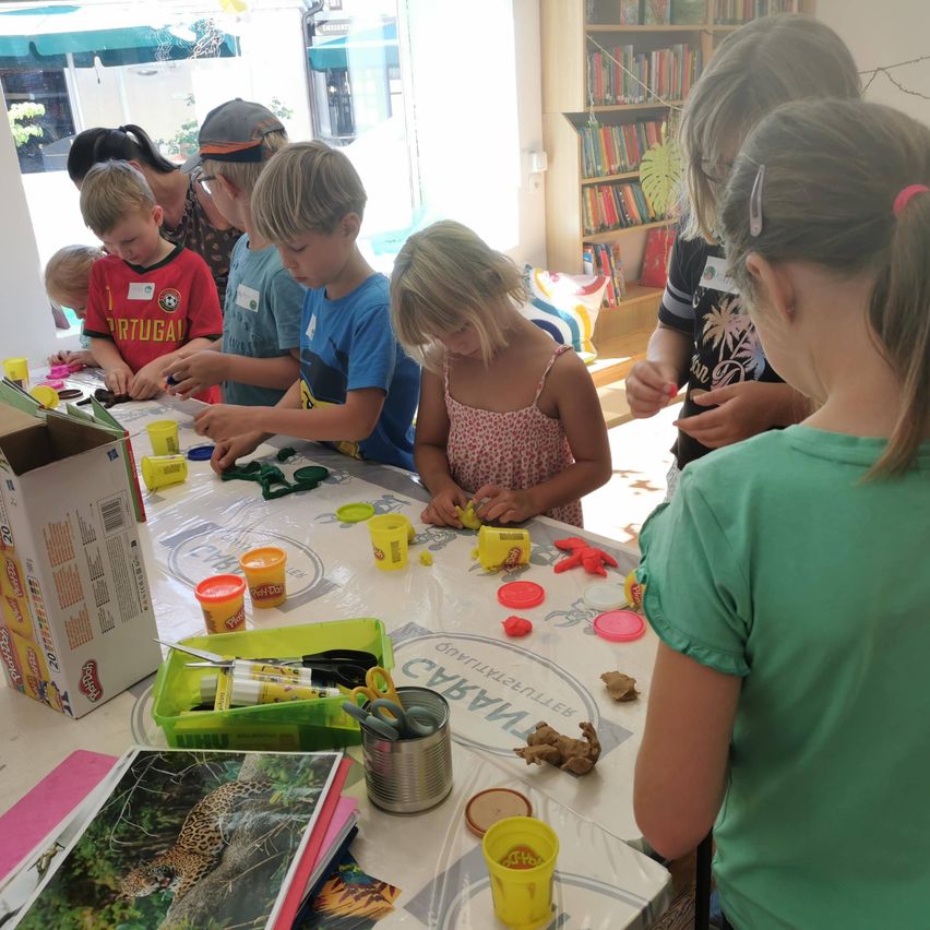 Kinder versammeln sich um einen Tisch und sind in einer Bastelaktivität mit verschiedenen farbigen Play-Doh und Werkzeugen beschäftigt. Ein Regal mit Büchern ist im Hintergrund sichtbar.