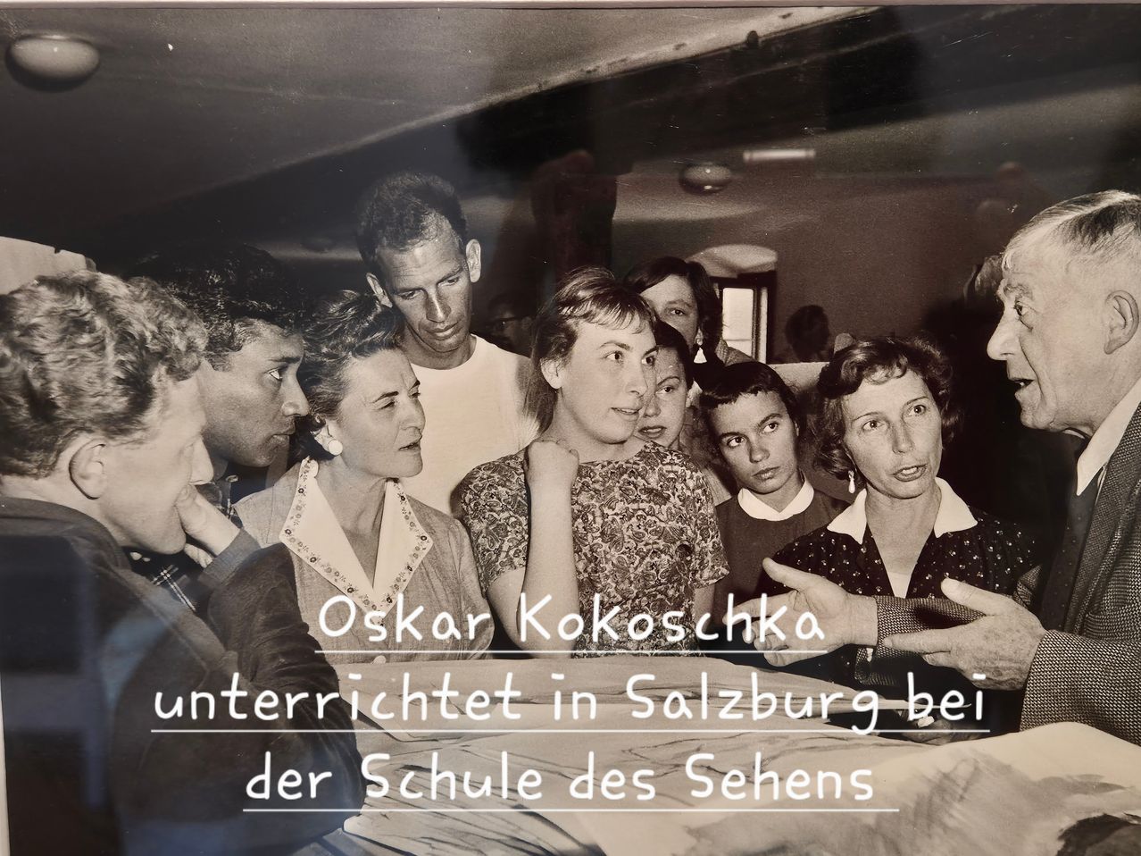 Ein Schwarz-Weiß-Foto von Oskar Kokoschka, der in Salzburg an einer Schule unterrichtet. Mehrere Schüler sind aufmerksam um ihn herum versammelt. Kokoschka zeigt auf ein Blatt Papier auf einem Tisch.
