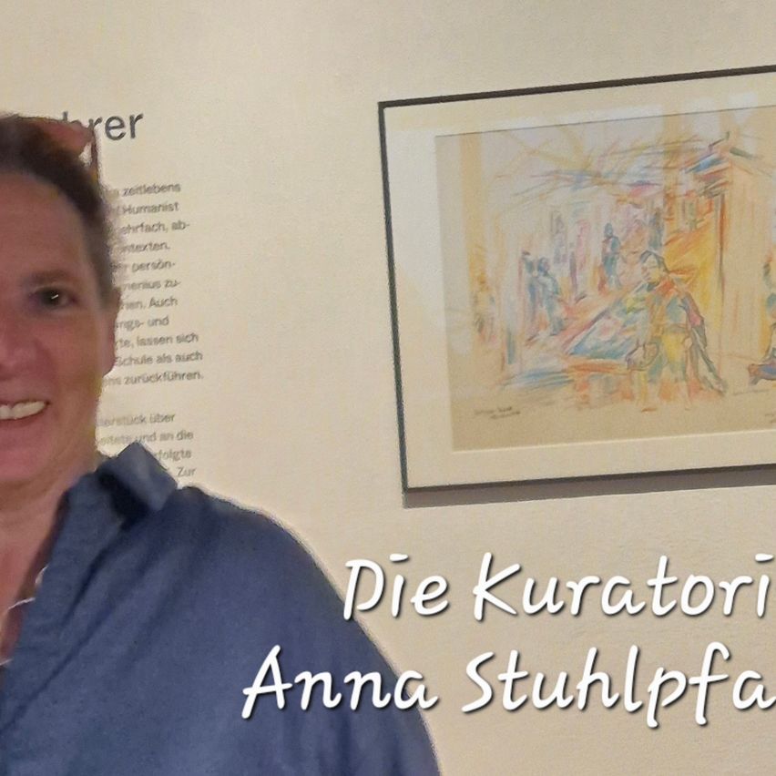 Eine lächelnde Frau steht vor einem Bilderrahmen mit einem lebhaften, abstrakten Gemälde von Menschen in einer Stadt. Die Wand hinter ihr ist weiß mit darauf geschriebenem Text. Die Frau trägt ein blaues Hemd und hat eine Brille auf dem Kopf.