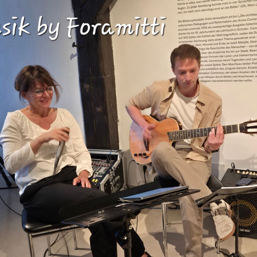 Zwei Musikerinnen führen einen Auftritt im Inneren durch. Eine Frau mit Brille hält ein Mikrofon, und ein Mann spielt Gitarre. Sie sitzen vor einer weißen Wand mit deutschem Text.