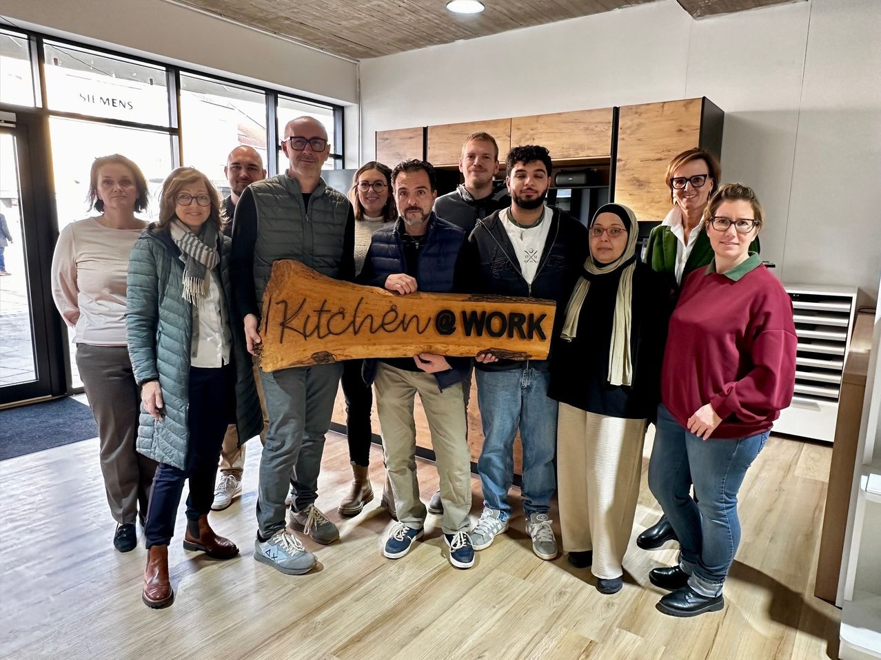 Eine Gruppe von Menschen steht in einem Raum mit Holzböden und hält ein Holzschild mit der Aufschrift Kitchen@Work. Sie sind in Winterkleidung gekleidet. Hinter ihnen ist ein Schrank mit Glasfenstern zu sehen.