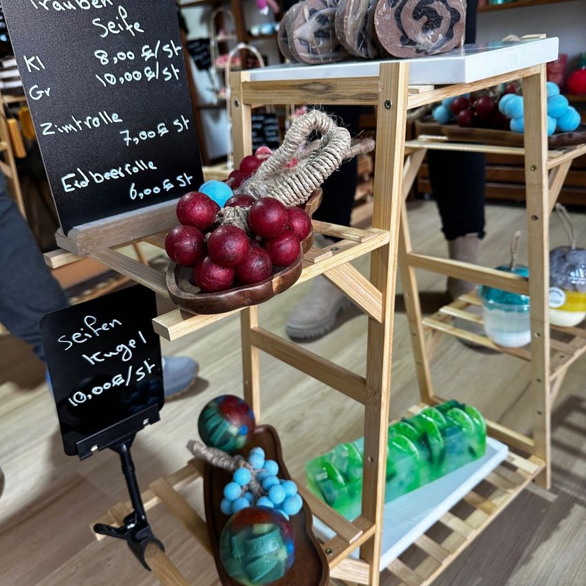 Ein Holzregal zeigt verschiedene Artikel. Eine Schüssel enthält rote Seifenkugeln mit einem Seil. Eine weitere Schüssel hat grüne Seifenblöcke. Ein schwarzes Brett zeigt die Preise für verschiedene Artikel.