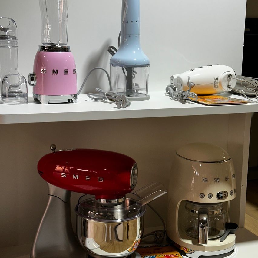 Ein Küchenregal zeigt verschiedene Smeg-Geräte, darunter einen roten Mixer, einen rosa Mixer, einen blauen Zerkleinerer und ein weißes Gerät mit Kabel.