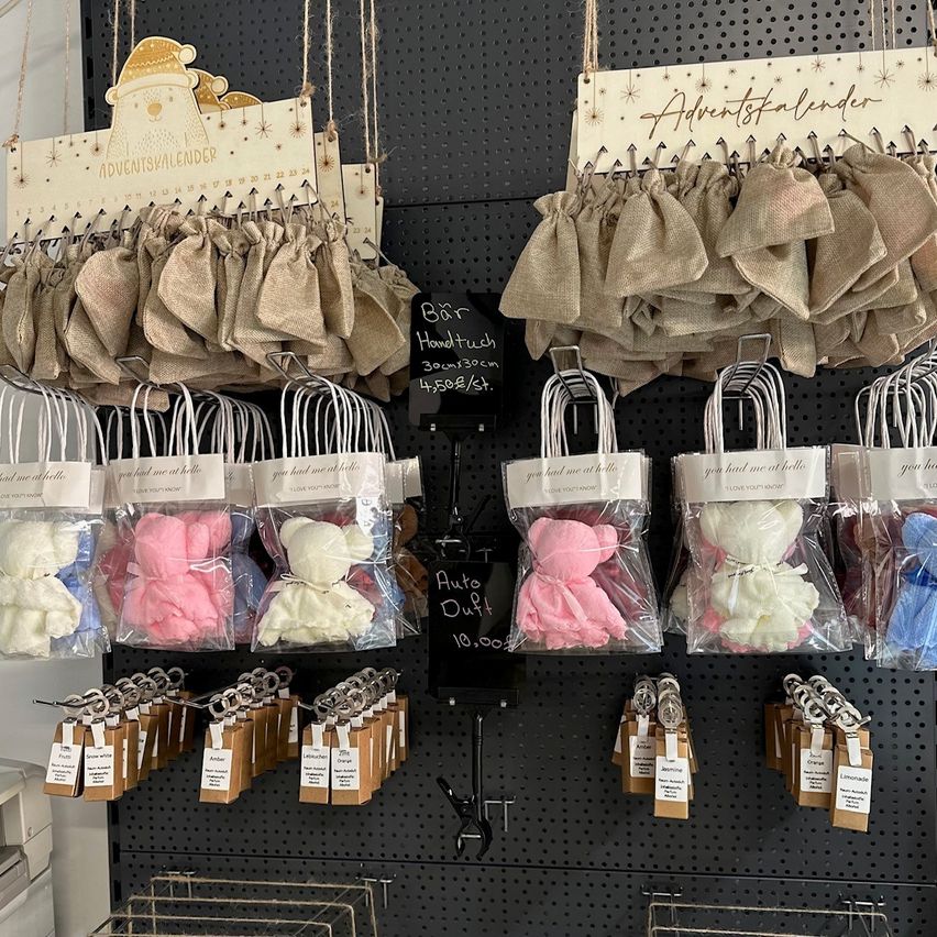 Eine Vielzahl von dekorativen Artikeln für die Feiertage sind ausgestellt. Es gibt braune Jutebeutel, rosa und weiße Badebomben in Plastikverpackungen und kleine Beutel, die an Haken hängen.