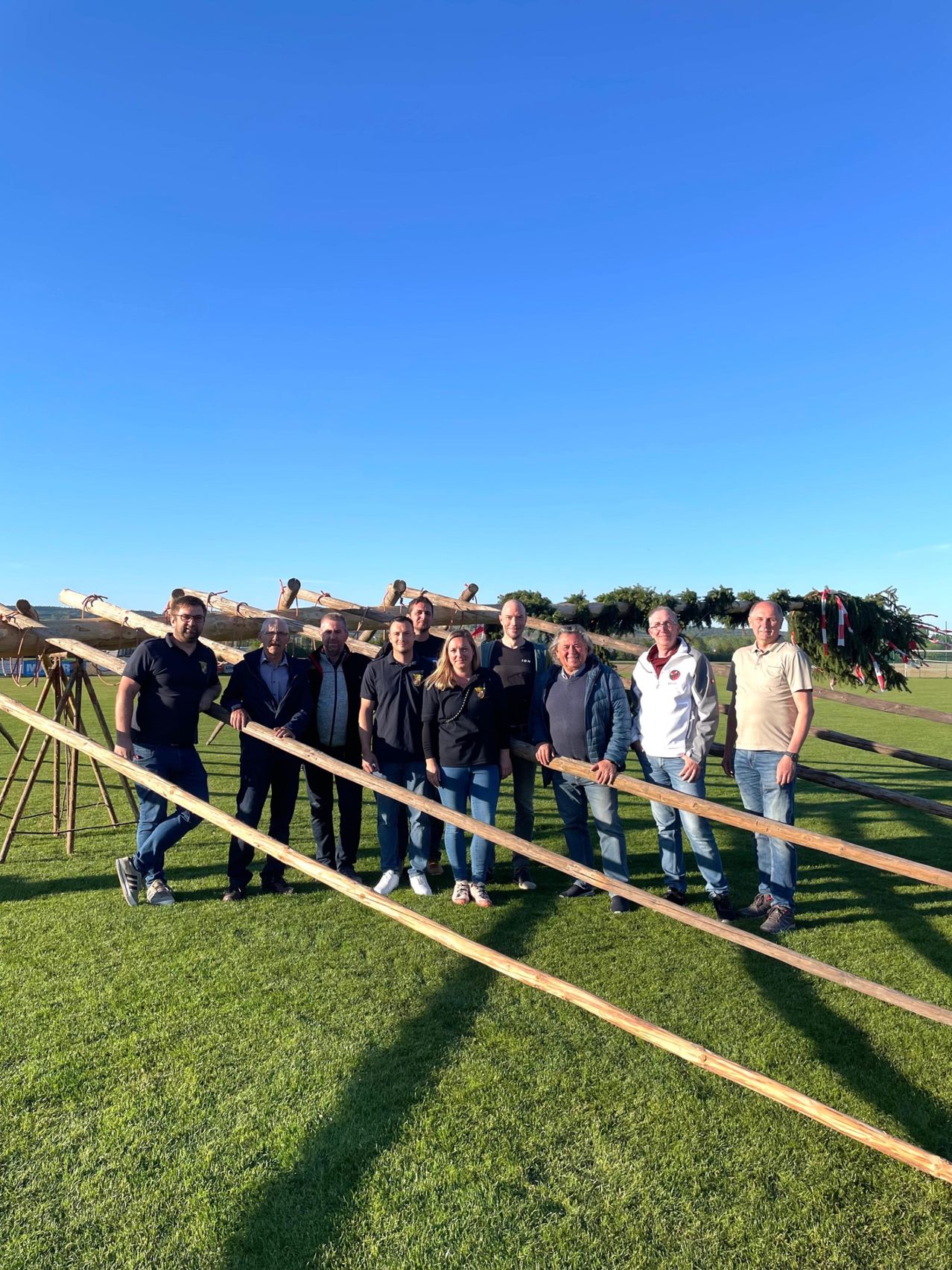 Eine Gruppe von Menschen in passender Kleidung posiert für ein Foto vor hölzernen Barrieren und Grünpflanzen an einem sonnigen Tag.