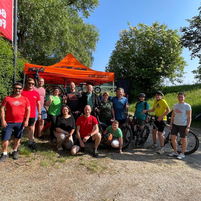 Eine Gruppe von Menschen in Fahrradausrüstung steht vor einem KTM-Zelt, einige mit Helmen und Fahrrädern, mit einem Grasbereich und Bäumen im Hintergrund.