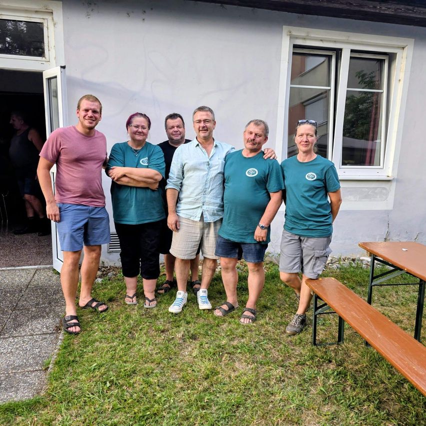 Fünf Menschen stehen vor einem weißen Gebäude und lächeln für ein Foto. Sie tragen Freizeitkleidung und einige tragen Brillen. Hinter ihnen steht ein Holztisch und eine Bank.