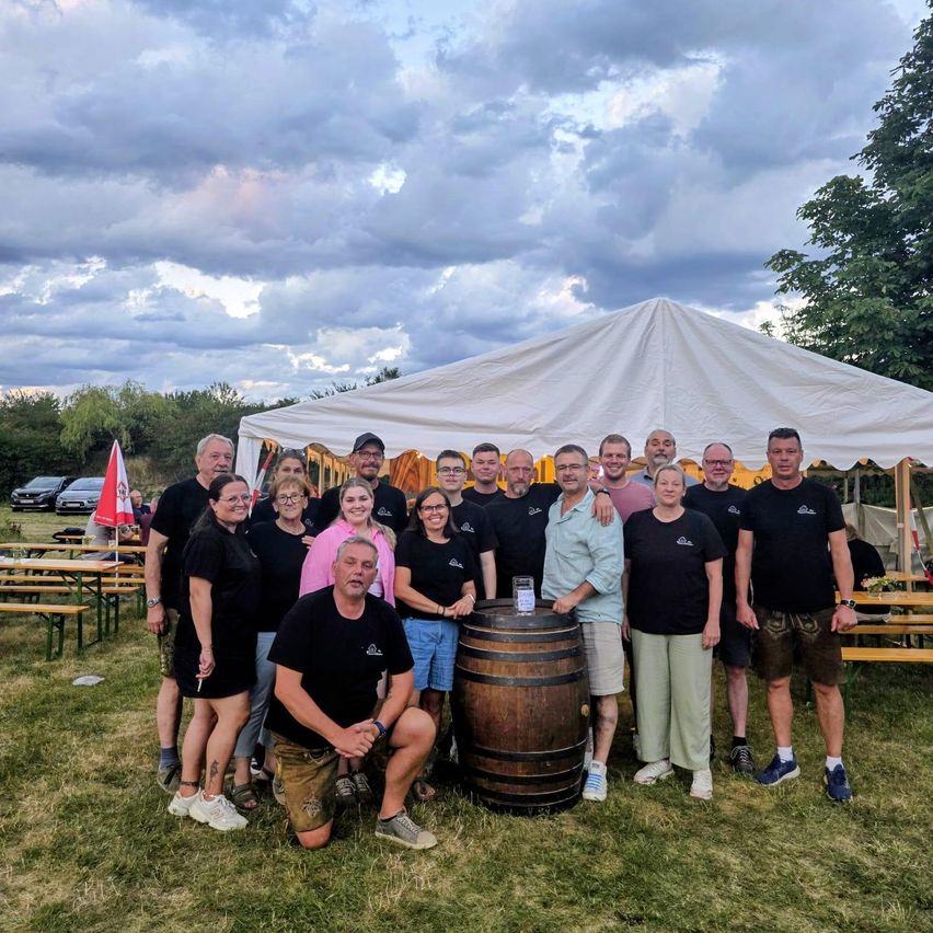 Eine Gruppe von Menschen in passenden schwarzen T-Shirts posiert für ein Foto auf einem Grasfeld mit einem Fass und einem Zelt im Hintergrund.
