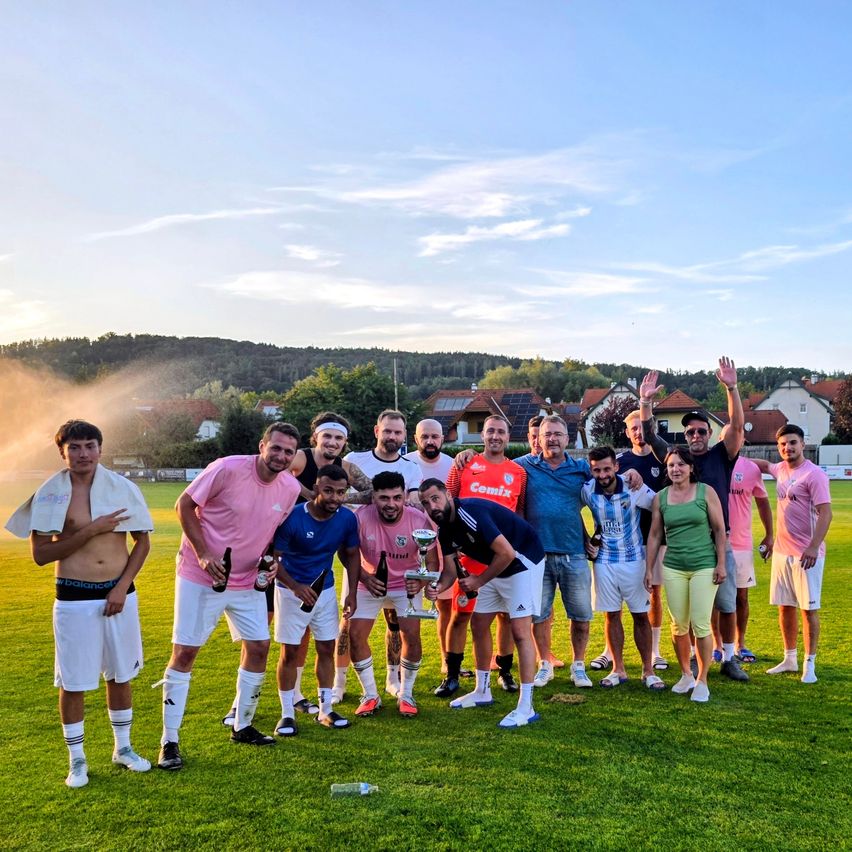 Eine Gruppe von Menschen, möglicherweise ein Sportteam, posiert für ein Foto auf einem Rasenfeld mit klarem Himmel und Häusern im Hintergrund. Einige halten Flaschen und einen Pokal.