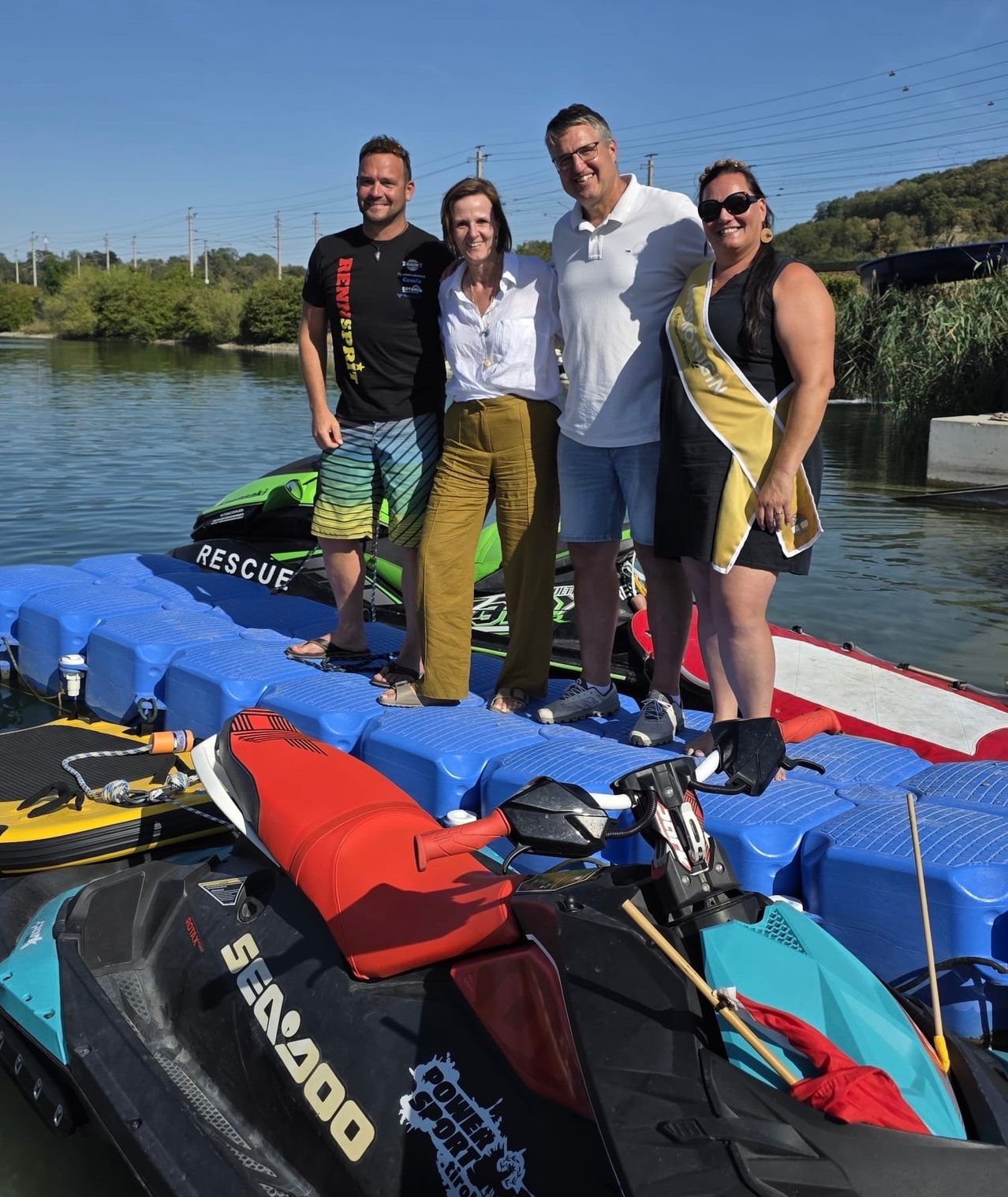 Vier Personen stehen auf einer schwimmenden Plattform auf einem See, mit zwei Jetskis im Hintergrund. Sie lächeln und posieren für ein Foto.