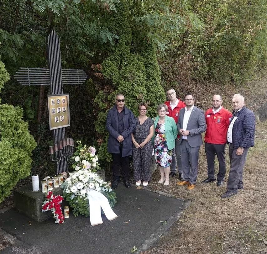 Eine Gruppe von Menschen steht um ein Denkmal mit einem Kreuz, Blumen und Kerzen, gekleidet in formeller Kleidung.