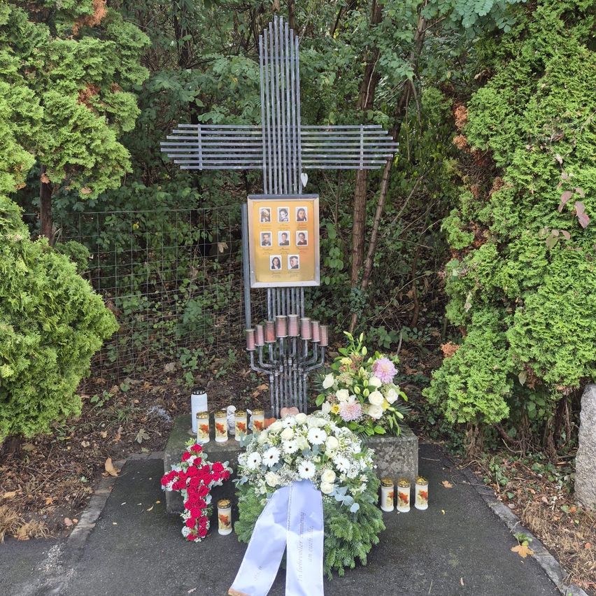 Ein Gedenkkreuz mit Fotorahmen und Blumen, umgeben von Kerzen und einem Band, in einer Gartenumgebung.