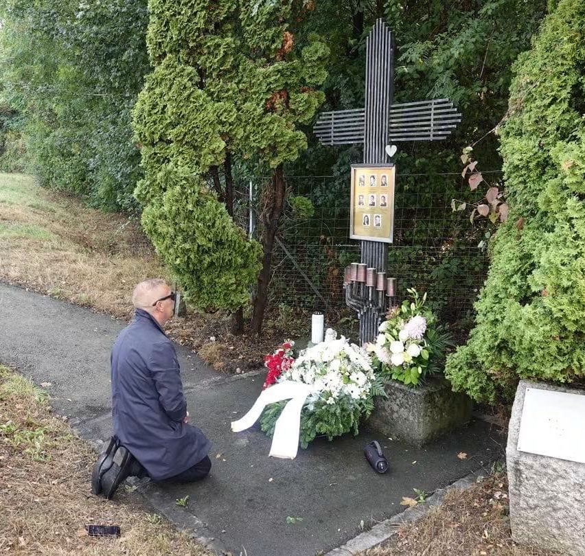 Ein Mann kniet vor einem Gedenkkreuz mit Fotoplakette und Blumen.