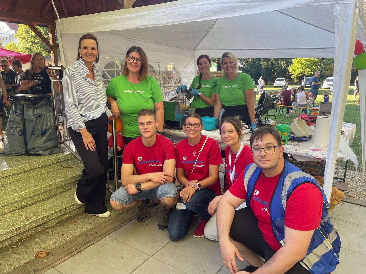 A group of volunteers in green and red shirts are posing for a photo under a tent at an outdoor event.