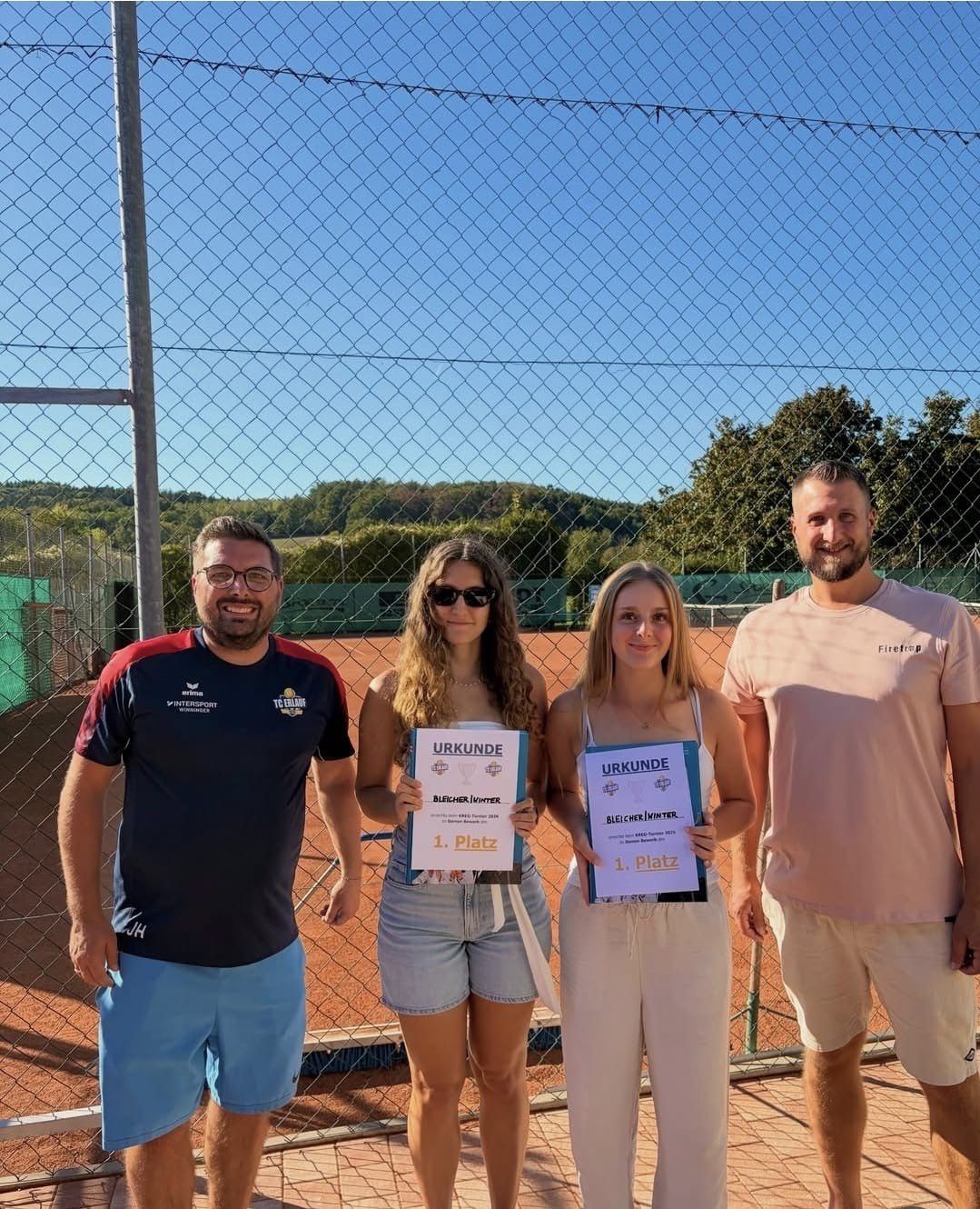 Four people stand on a tennis court, smiling and holding certificates. Two women hold blue certificates, and two men stand on either side. The background shows a fence and trees.