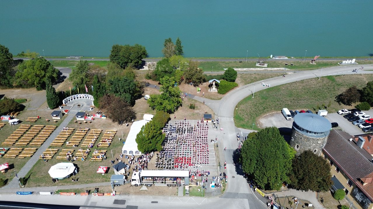 Aerial view of a large gathering near a lake, with people in red shirts, a white tent, and a white truck. Trees and benches are scattered around.