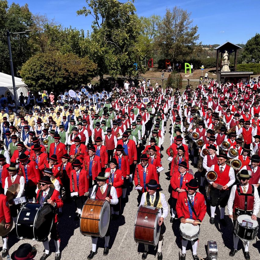 A large group of people dressed in red and green costumes and hats are marching in a parade, carrying drums and brass instruments. The setting is outdoors with trees and a small structure in the background.