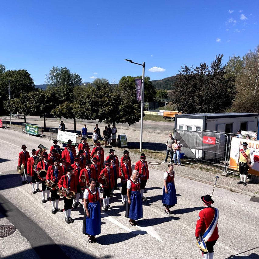 A marching band dressed in red and blue uniforms is performing on a street with trees and a clear sky in the background.