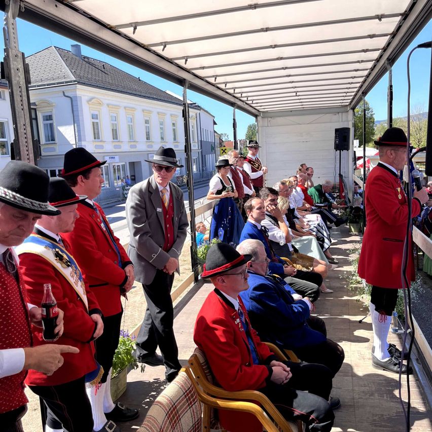 A group of men in red uniforms and hats are standing on a stage with a microphone. A man in a gray suit stands in front of them. Behind them, people are sitting on chairs, and a speaker is on the right. A building is in the background.