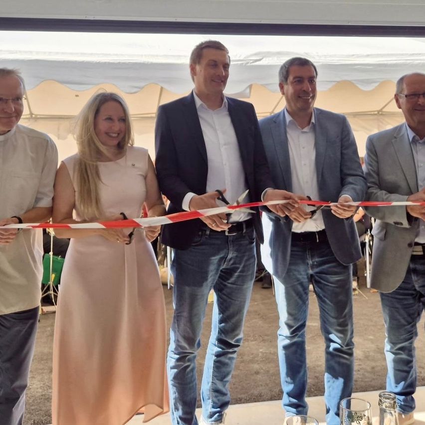 Five people stand under a tent, smiling, as they cut a ribbon with scissors. The woman on the left wears a pink dress, and the men wear suits.