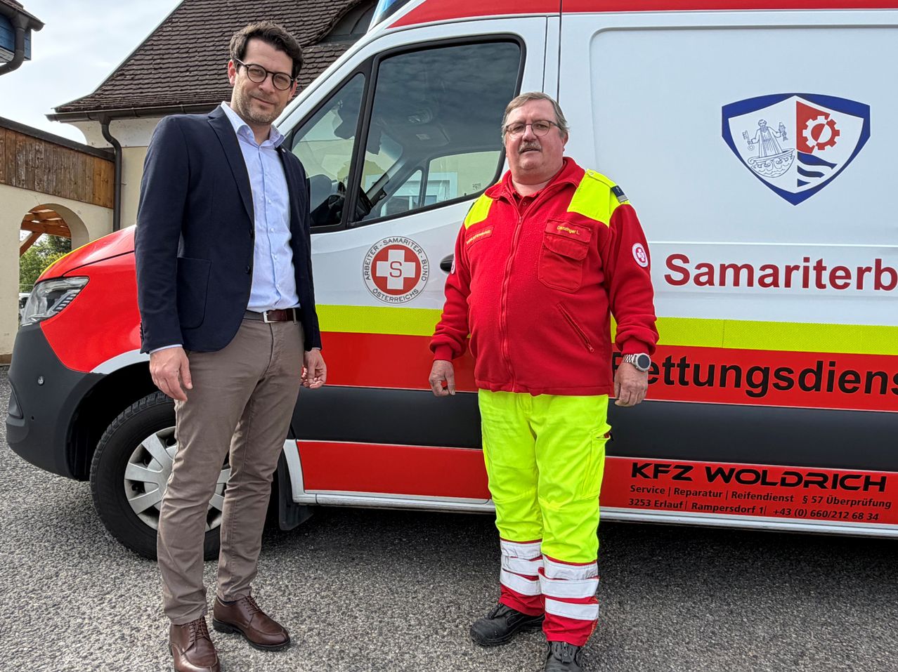 Zwei Männer stehen vor einem Krankenwagen mit dem Wort SAMARIT an der Seite. Der Mann auf der rechten Seite trägt eine rot-gelbe Jacke mit reflektierenden Streifen. Der Mann auf der linken Seite trägt einen blauen Blazer und khakifarbene Hosen. Sie schauen beide nach vorne.