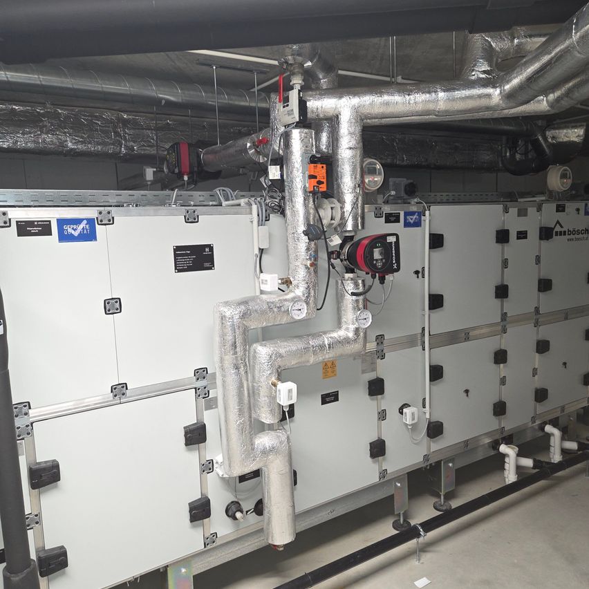 An industrial setting with a large white machine, multiple pipes, and various gauges and meters. The machine has a blue label with text and a logo.